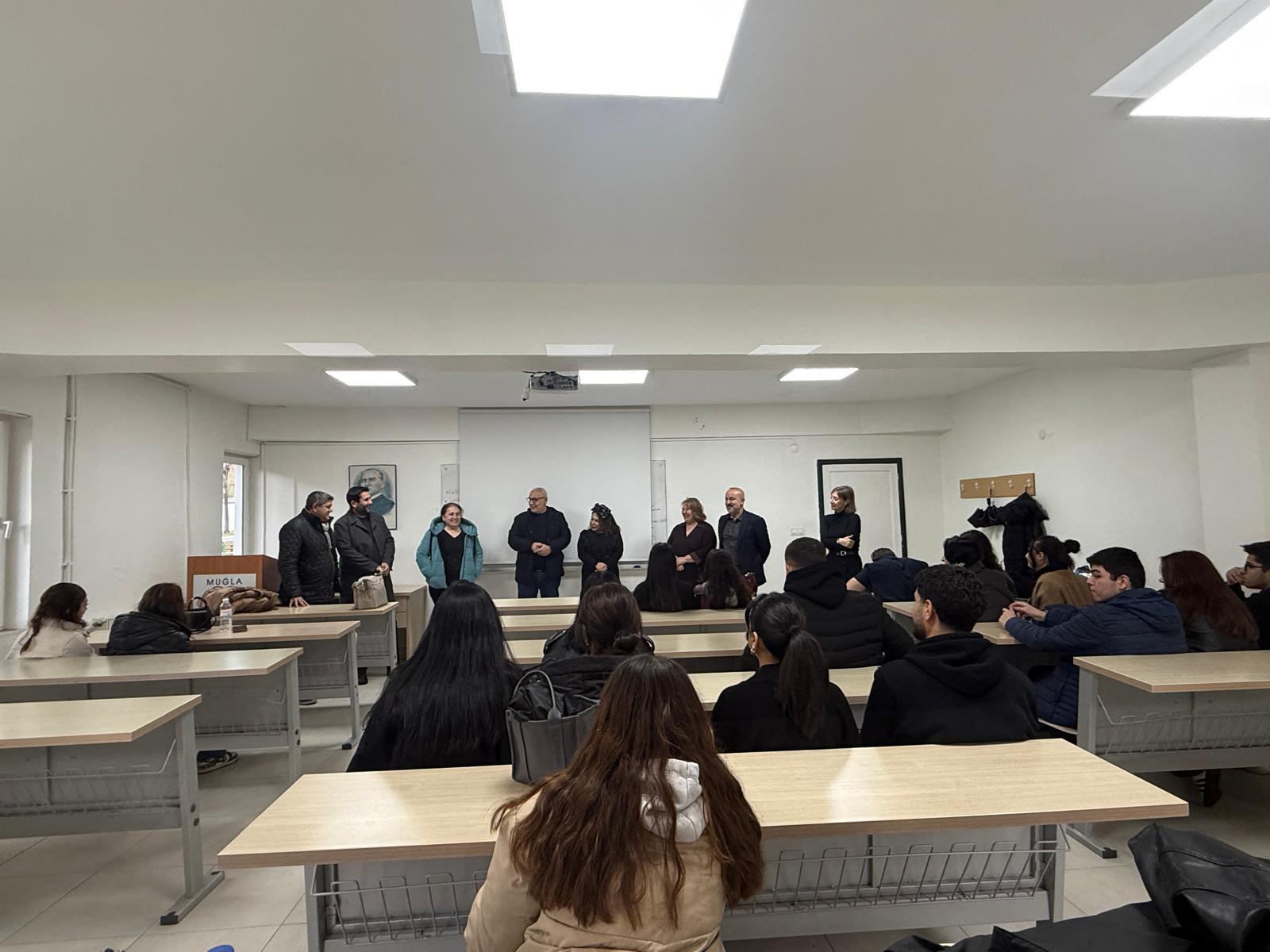Bir sınıfta ve tahtada kadın ve erkeklerden oluşan yaklaşık 10 kişi öğrenciler ile gülümseyerek konuşuyor. 